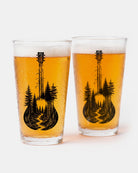 Two clear drinking glasses filled with golden beer and foam, each printed with a black silhouette of a guitar shaped like a pine forest, on a clean white background.