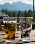 Two glasses with scenic designs on a wooden surface with a mountainous background